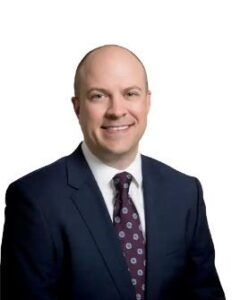 A smiling bald man in a suit and patterned tie poses for a portrait against a white background.