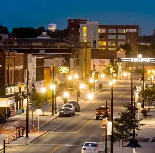 A well-lit, city street at dusk featuring parked cars, small businesses, and a visible “Broadway” sign in the background, with a water tower seen in the distance.