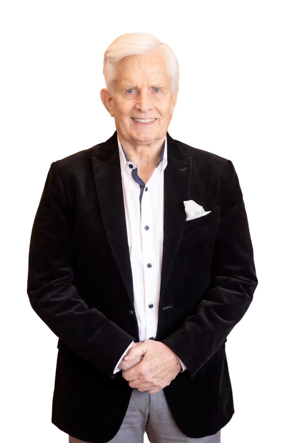 Elderly man with white hair in a black blazer, white shirt, and gray pants, standing with hands clasped.